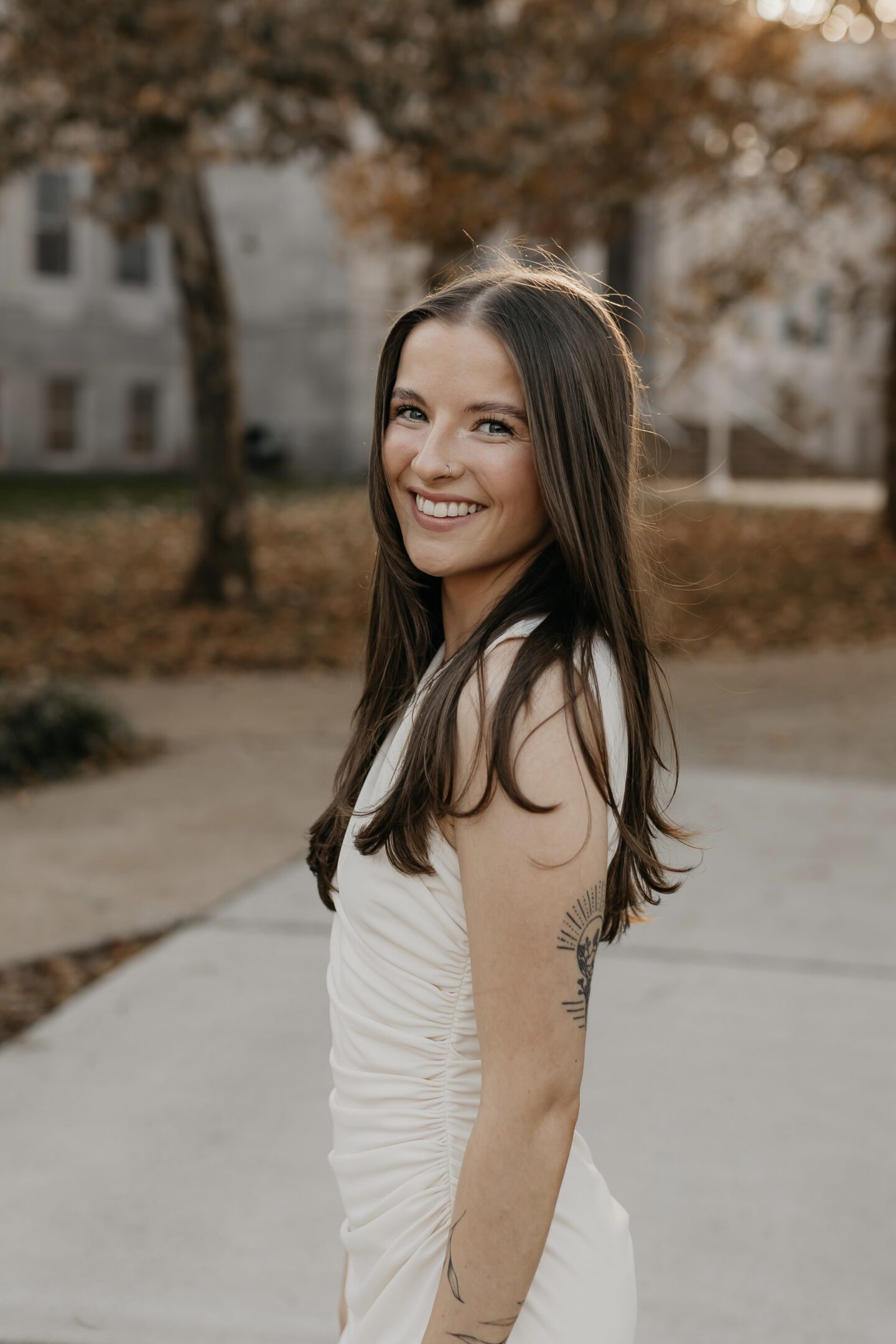 Smiling woman with long brown hair in a white sleeveless top outdoors.