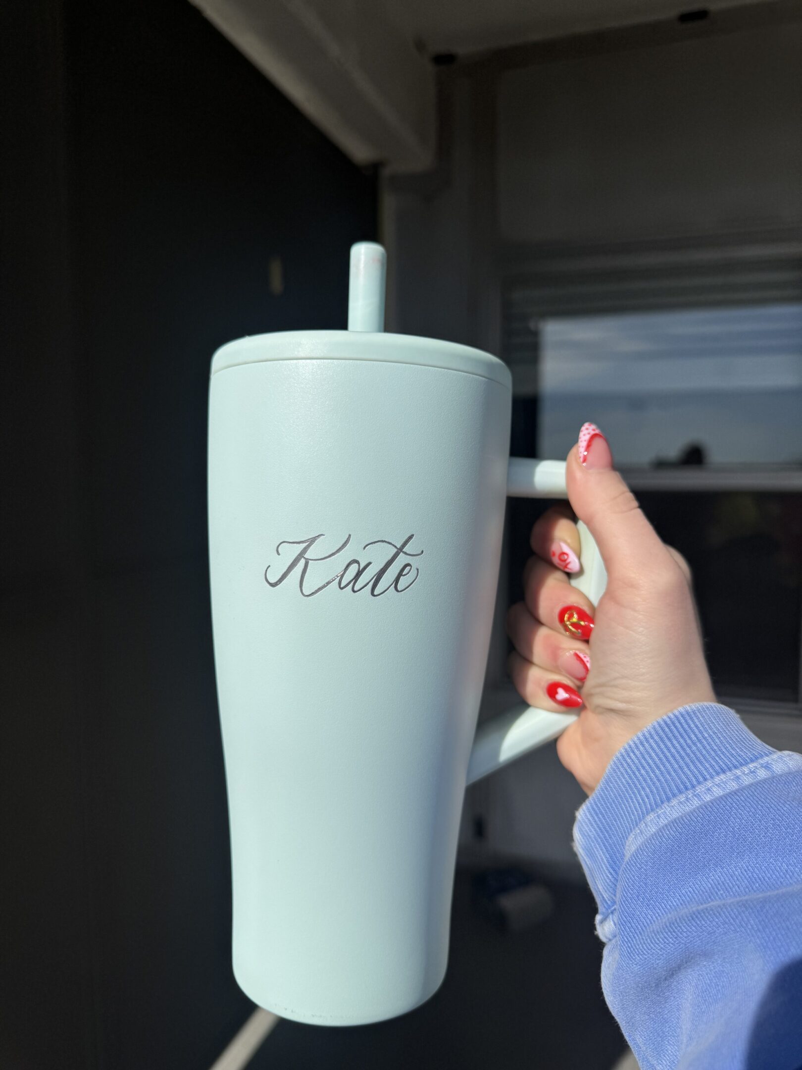 White tumbler with the name 'Katie' engraved, held by a hand with decorated nails.