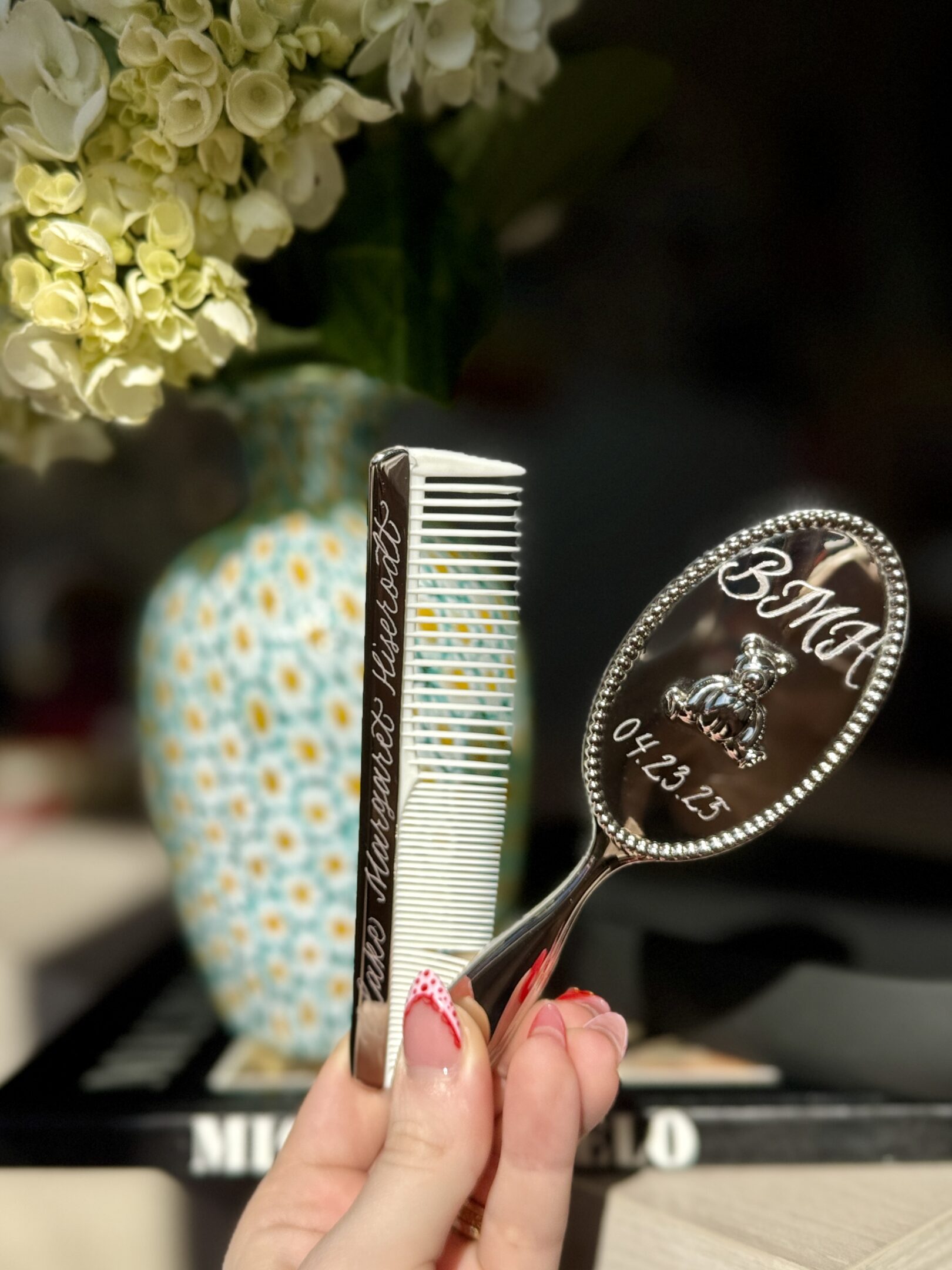 A hand holding a personalized mirror and a comb near a decorative vase.
