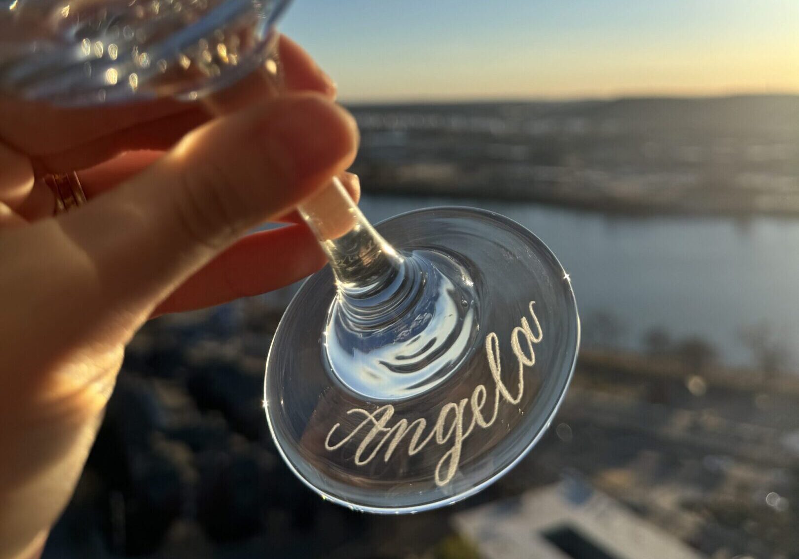 A hand holding a wine glass engraved with the name Angela during sunset.